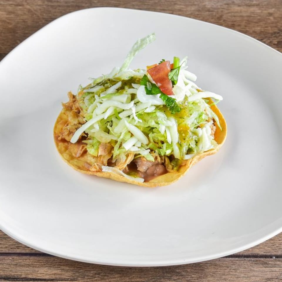A single Vaqueros Mexican Restaurant tostada, topped with shredded lettuce, salsa verde, and diced tomatoes.