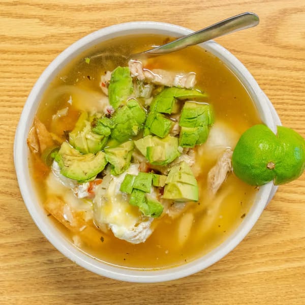 Overhead view of a bowl of Vaqueros Mexican Restaurant's chicken soup with avocado, cheese, and a lime.